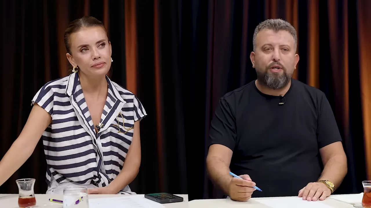 Sunucu sağda el hareketiyle konuşuyor, solunda eşlik eden kişi dinliyor; arka plan sade perde.