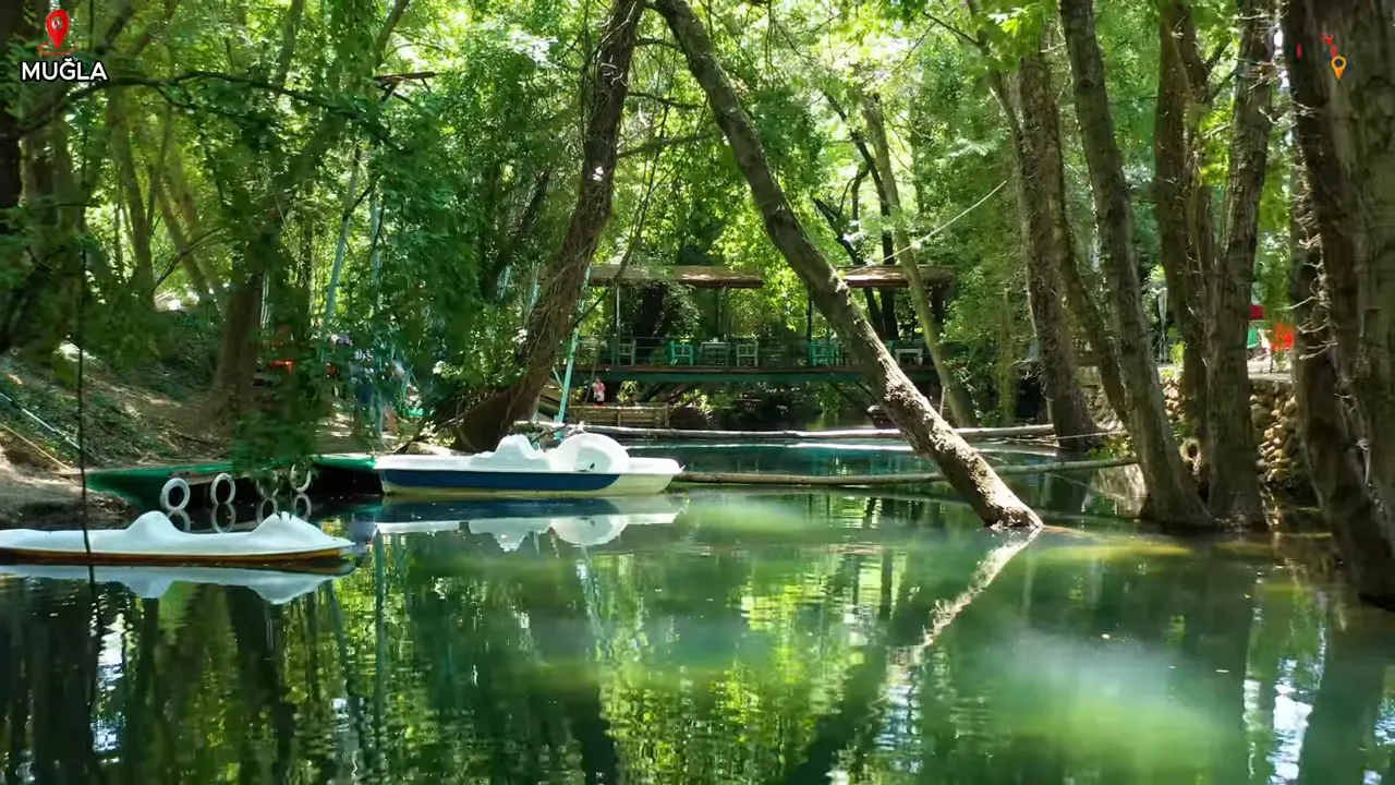 Ağaçlarla çevrili nehir, yüzen pedal tekneler ve arka planda pergola/restoran