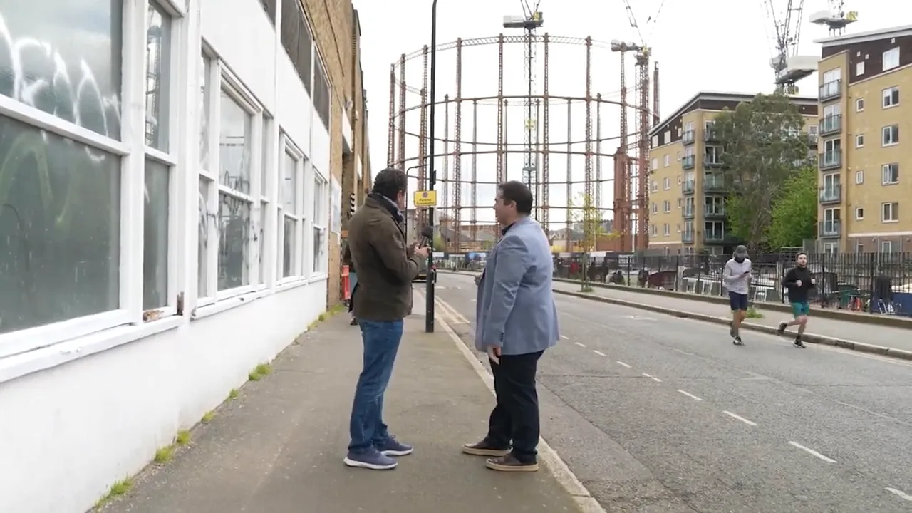 Arka planda Viktorya d&ouml;nemi gaz tankı iskeleti g&ouml;r&uuml;len sokakta konuşan iki kişi