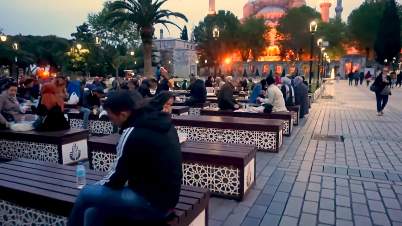 Rows of outdoor benches filled with people having iftar in front of a lit mosque at dusk.