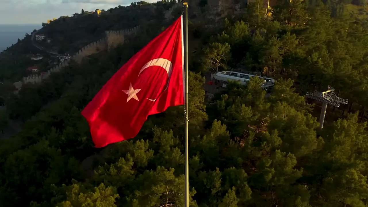 Turkish flag waving in front of a forested hillside with medieval coastal fortress walls.