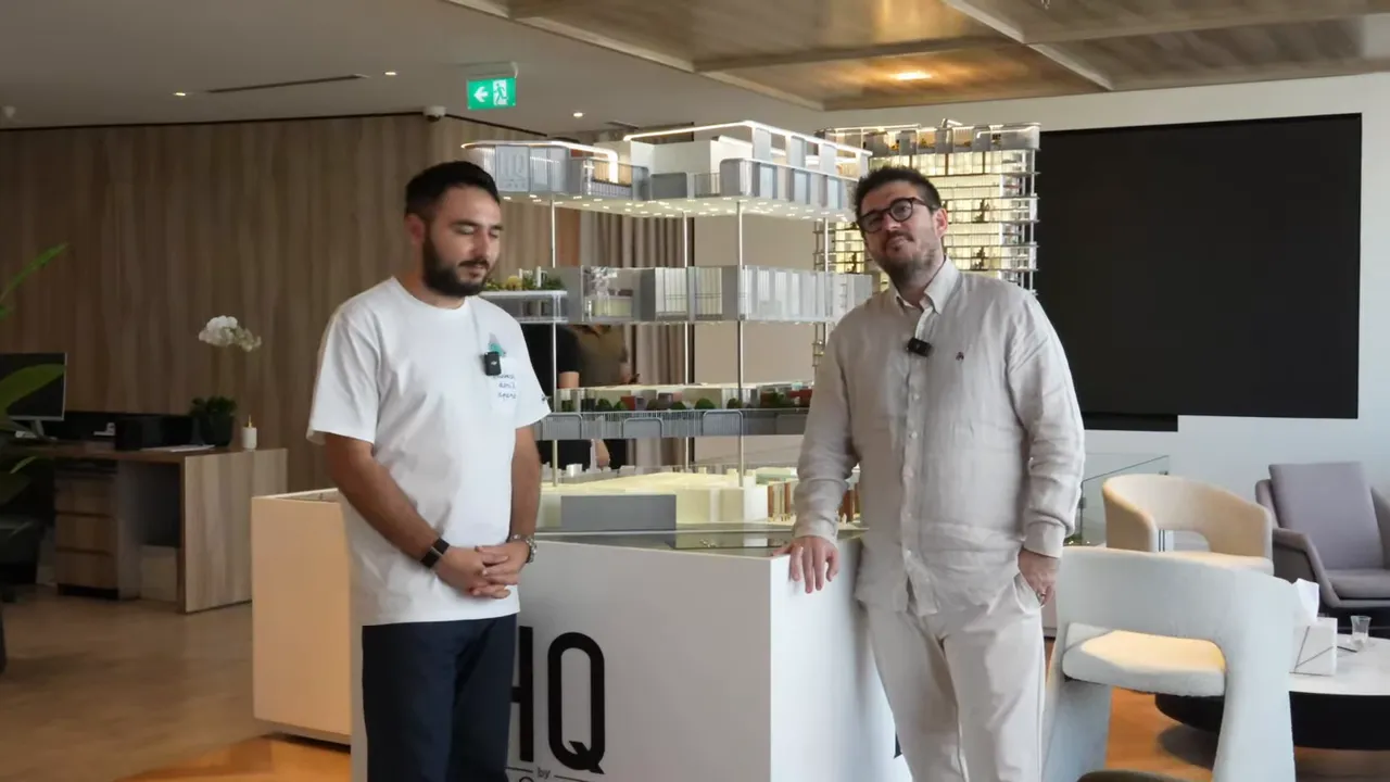 İki konuşmacı ofis proje maketi &ouml;n&uuml;nde duruyor; maket ve ofis i&ccedil; mekanı net g&ouml;r&uuml;n&uuml;yor.