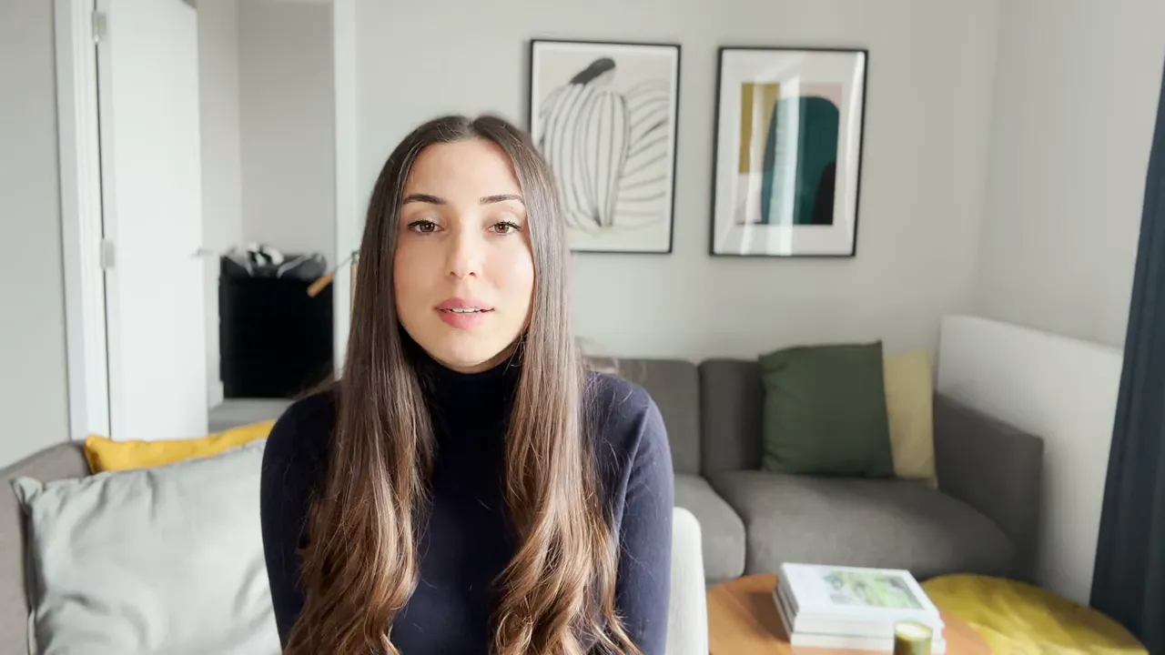 Sunucu oturma odasında doğrudan kameraya bakıyor, temiz ve net arka plan.