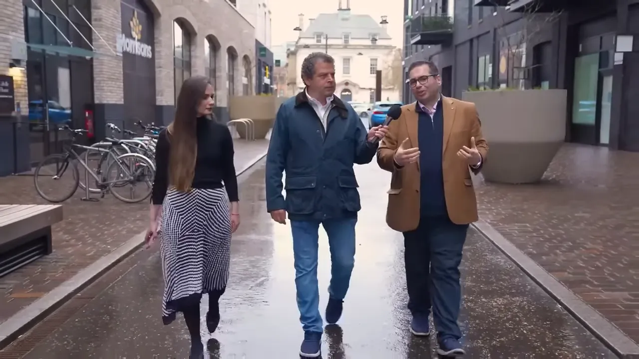 Brentford benzeri bir caddede &uuml;&ccedil; kişi y&uuml;r&uuml;rken, ortadaki sunucu mikrofona bakıyor; arka planda d&uuml;kkan cephesi ve bisiklet parkı g&ouml;r&uuml;l&uuml;yor.