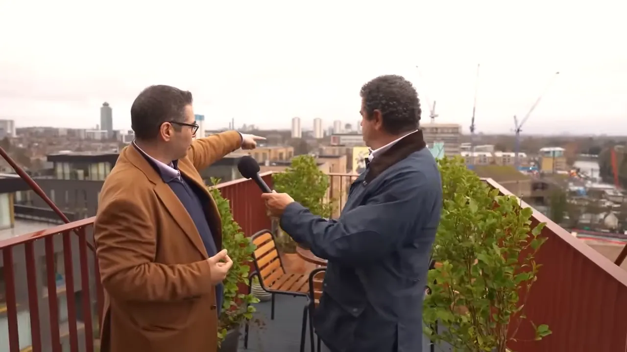 Terasta bulunan kişi projeyi parmağıyla işaret ediyor; yanındaki sunucu mikrofon tutuyor ve arka planda inşaat alanı ile nehir g&ouml;r&uuml;n&uuml;r.