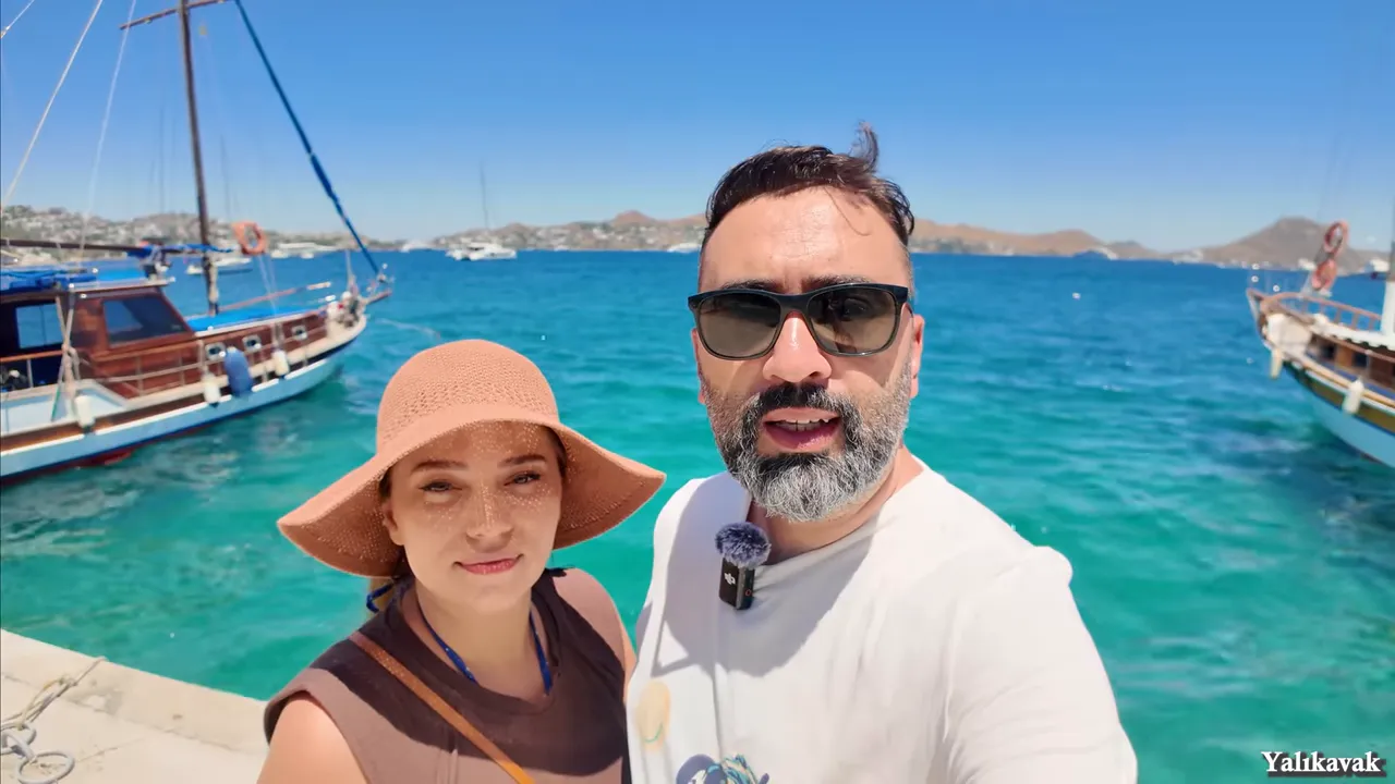 Yalıkavak iskelesinde guletler, turkuaz deniz ve iki gezginin ön planda olduğu görüntü