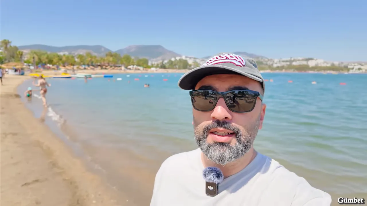 Gümbet plajının deniz kenarı, turkuaz su ve arka planda şezlonglarla plaj hattı, sunucu çerçeve kenarında