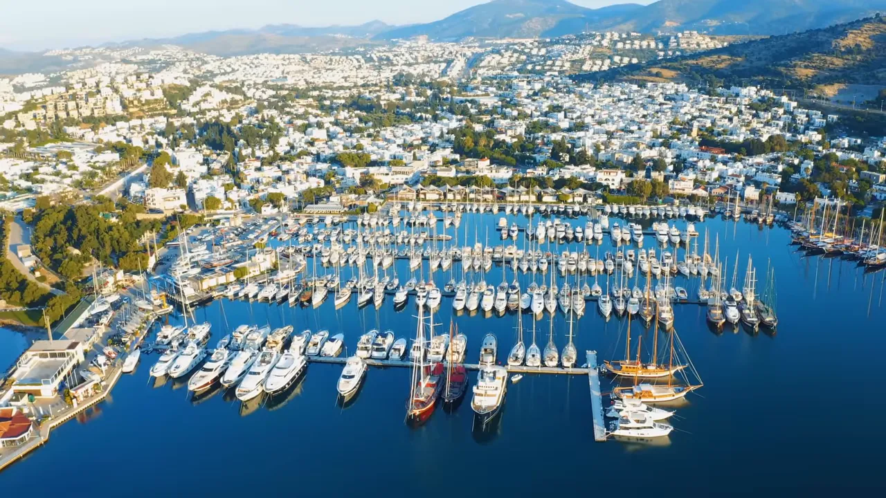 Aerial view of Bodrum marina with rows of yachts, clear blue water and white waterfront buildings.