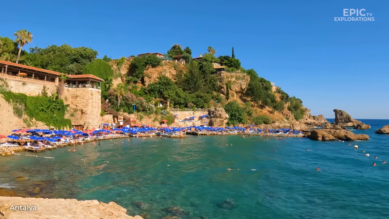 Wide view of an Antalya bay with clear turquoise water, sunbathers on platforms and rocky cliffs.