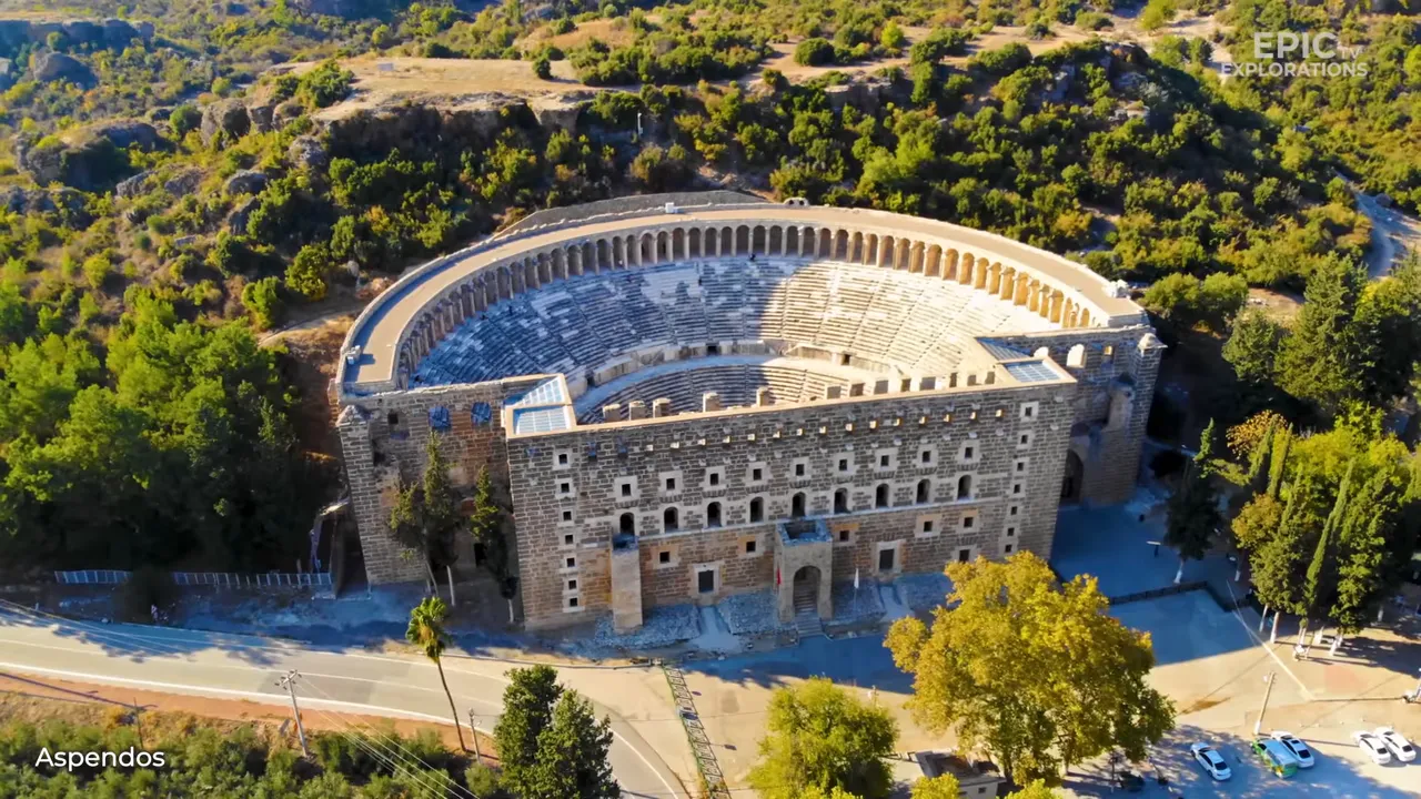 Wide aerial view of Aspendos Roman theatre with surrounding forest and road, showing the complete auditorium and stage.