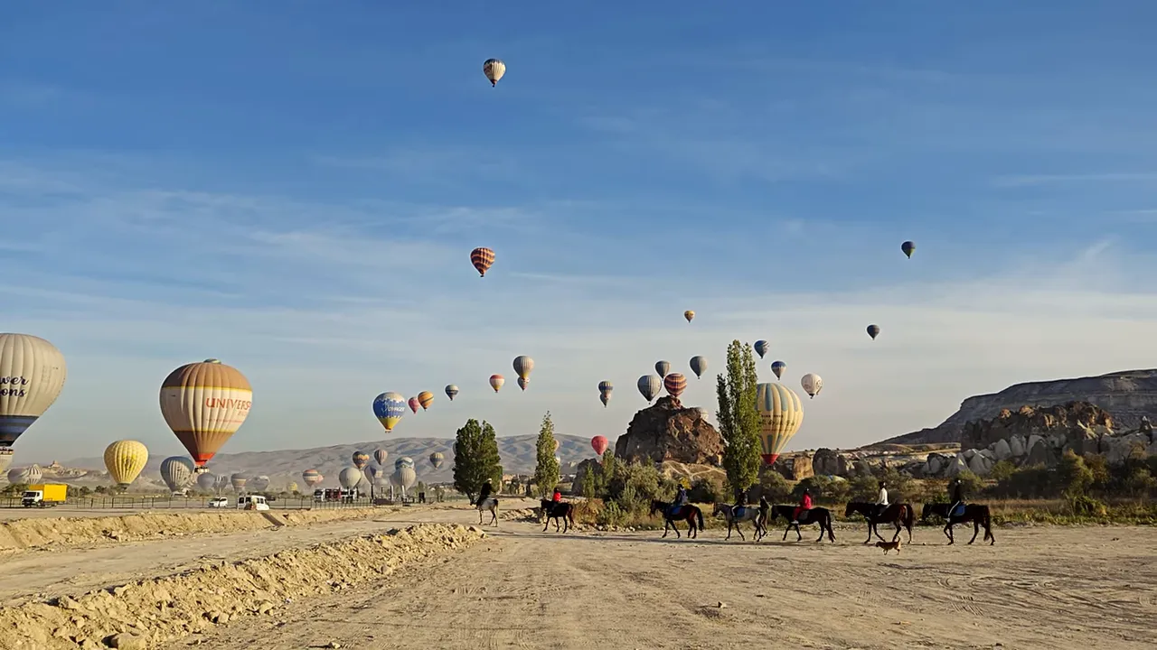 Numerous hot air balloons rising over Cappadocia with a line of people on horses in the foreground, clear blue sky.