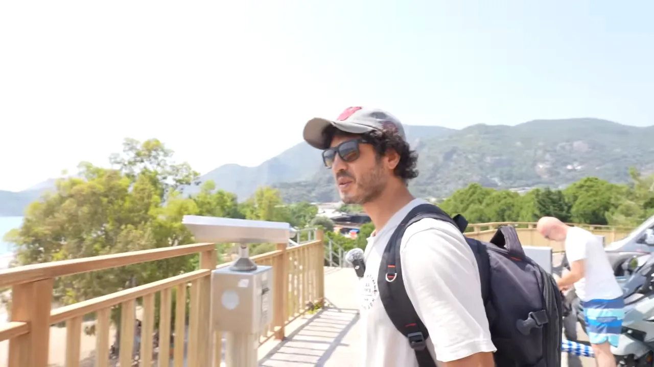 Deniz ve dağ manzarası ile tahta platform üzerinde yürüyen bir seyahatci; uzaklarda sahil ve teleskop.