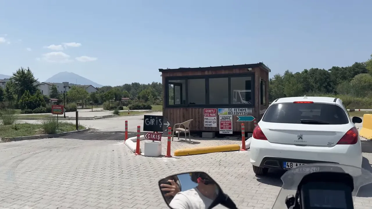 Giriş gişesi önünde bekleyen beyaz otomobil, gişe üzerinde ‘ücretli oto park’ ve belediye tabelaları.