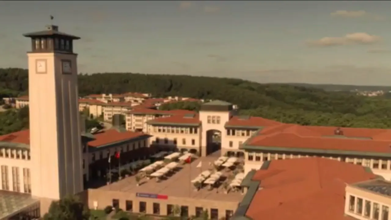 Koç University clock tower and central courtyard with tables and umbrellas, red-roofed buildings in the background