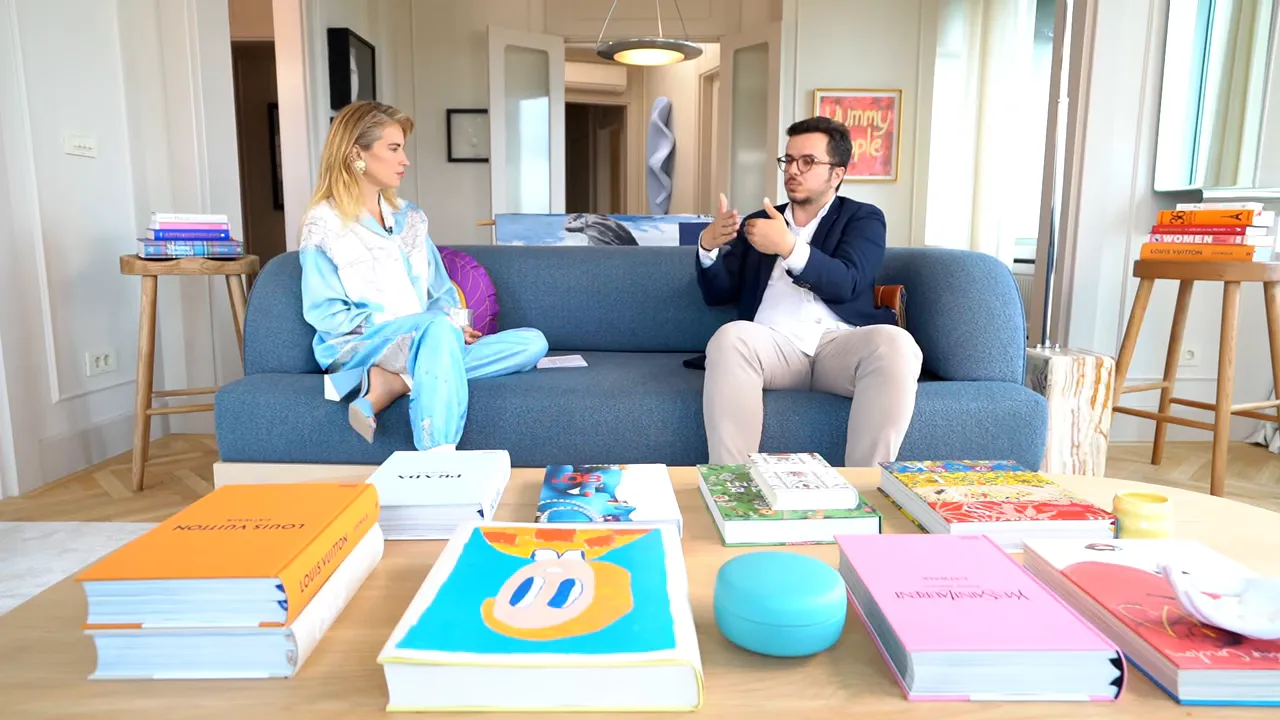 İki konuşmacı kanepe &uuml;zerinde birbirine bakıyor, &ouml;nde sehpa ve kitaplar net g&ouml;r&uuml;n&uuml;yor