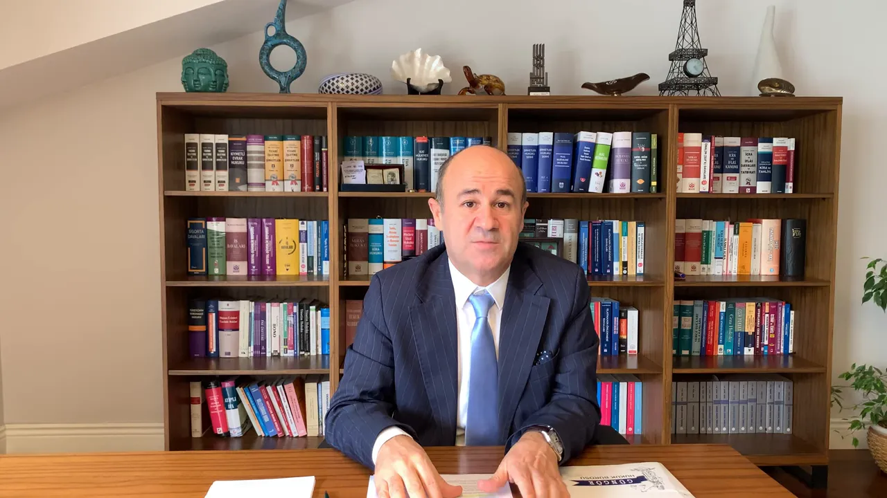 Net ve dengeli kadrajda masa başında oturan konuşan kişi; arka planda kitaplık