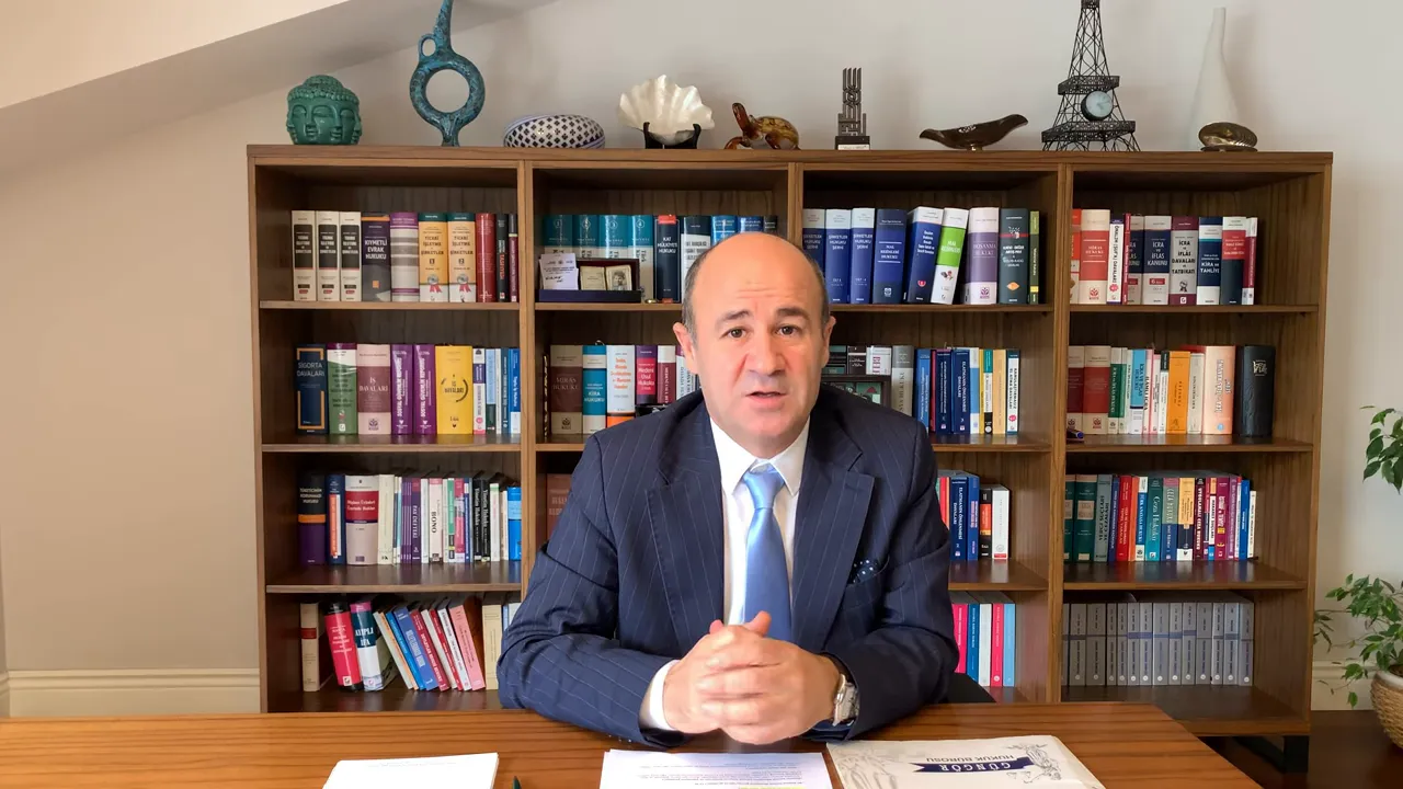 Ofis ortamında kitaplık &ouml;n&uuml;nde oturan konuşmacı, masa &uuml;zerinde başvuru belgeleri var.