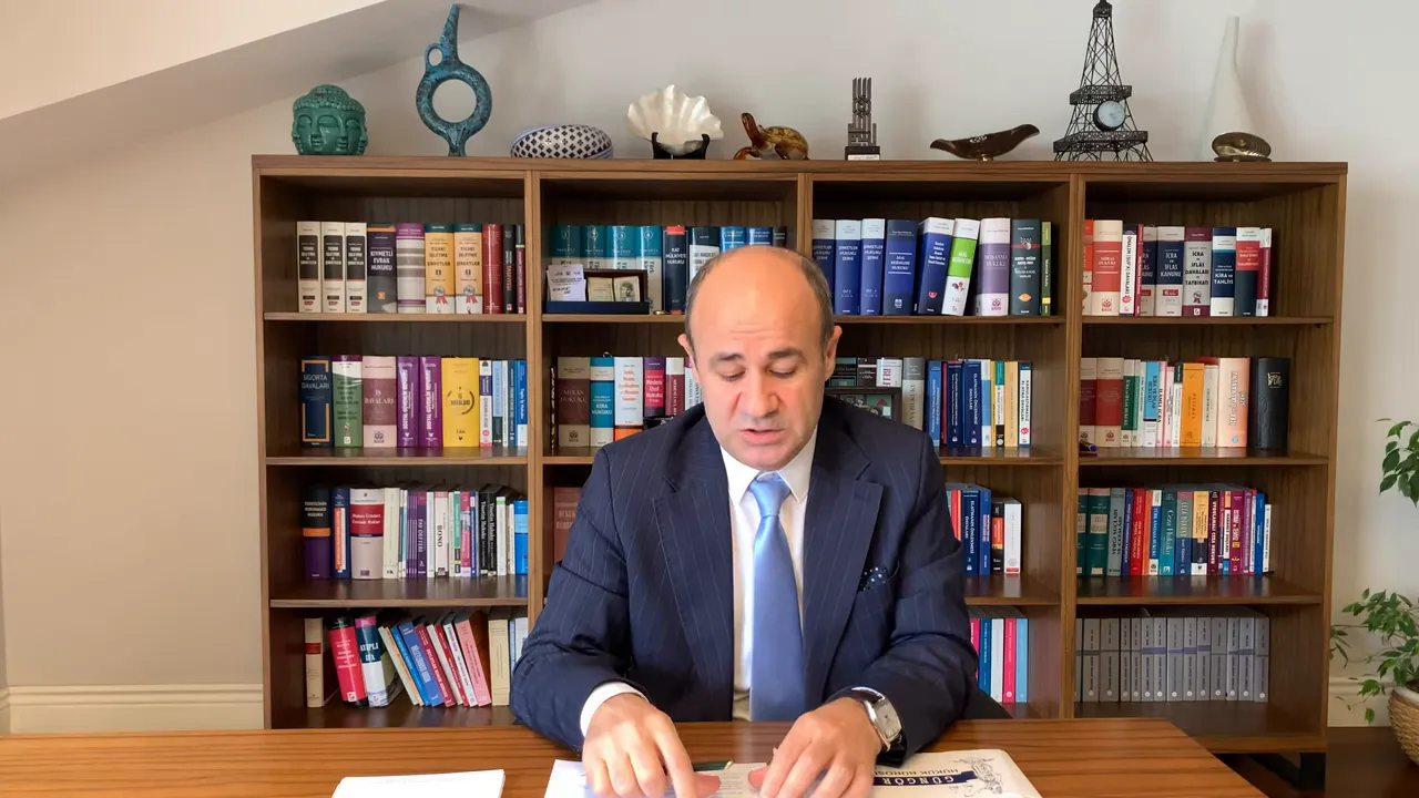 Konuşmacı masadaki belgeleri incelerken, arka planda dolu bir kitaplık g&ouml;r&uuml;n&uuml;yor.