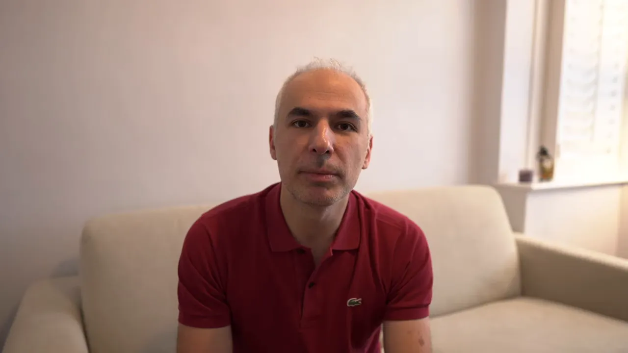 Konuşmacı kanepede merkezi olarak kadraja alınmış, iyi aydınlatılmış portre görüntüsü.