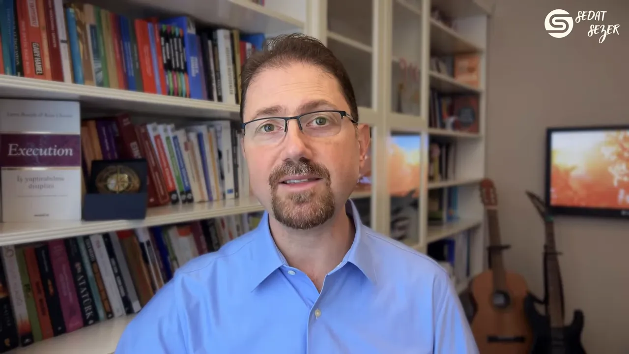 Konuşmacı ofis &ouml;n&uuml;nde direkt kameraya bakıyor; kitaplık ve gitar arka planda.