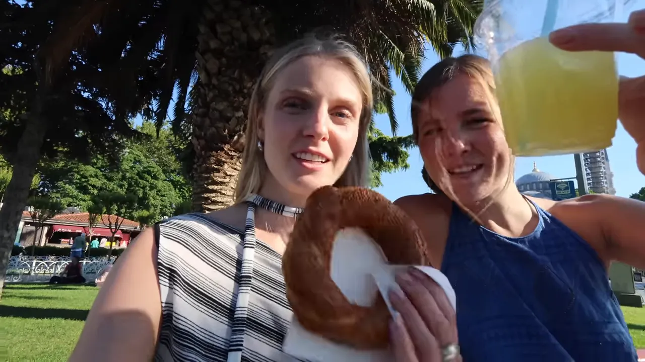 Two friends holding a sesame simit and a cup of fresh lemonade while sitting on a park bench.