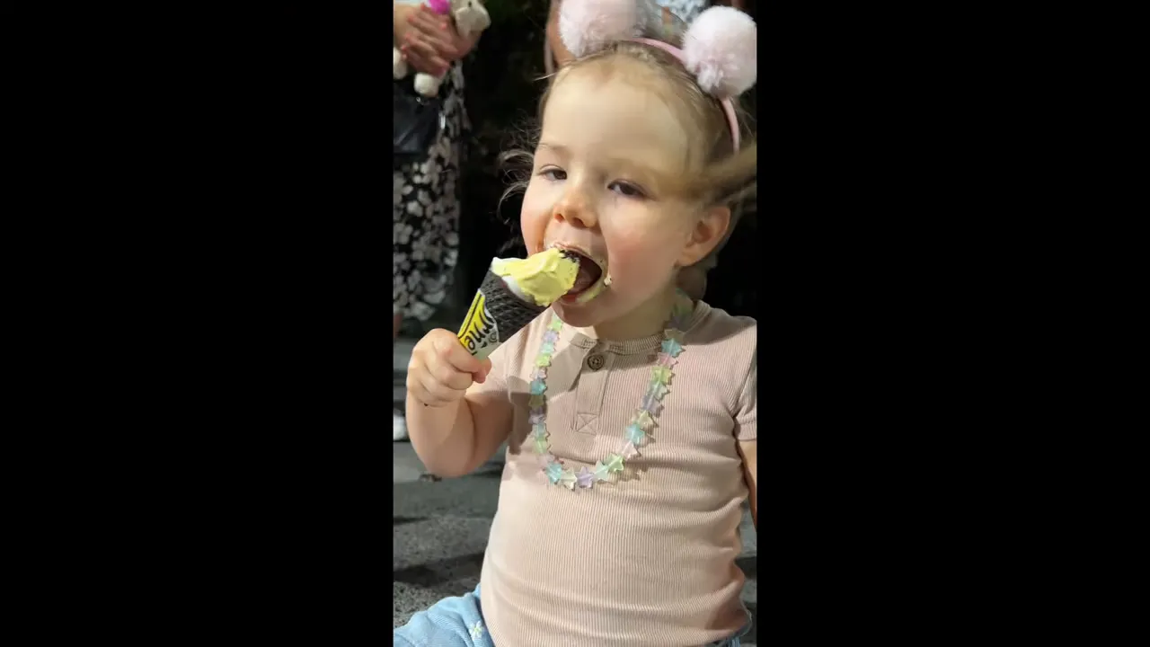 Toddler eating an ice cream cone outdoors, wearing a star necklace and fluffy headband