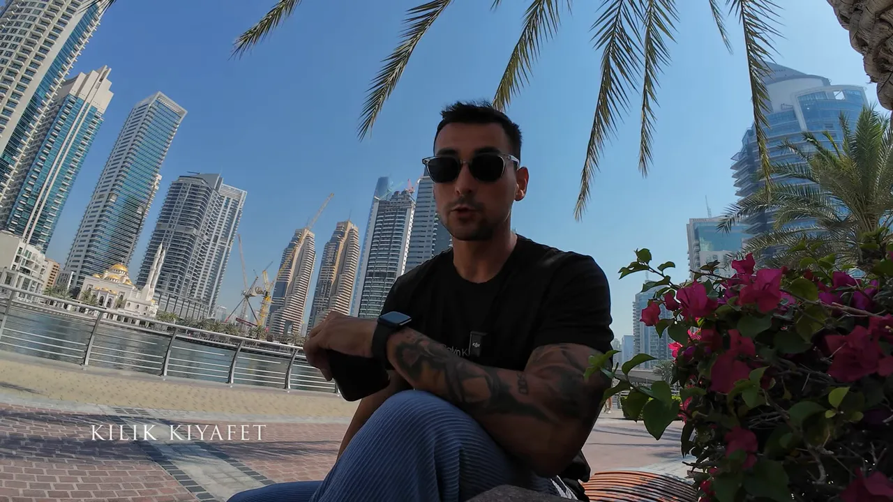 Marina &ouml;n&uuml;nde oturan bir kişi, arka planda Dubai g&ouml;kdelenleri ve ekranda 'KILIK KIYAFET' yazısı.