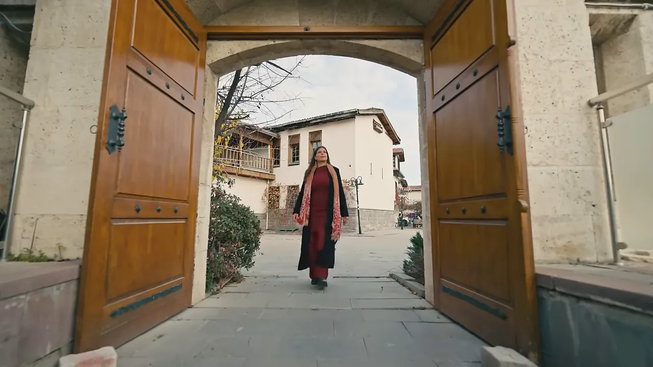Presenter walking through an open wooden gate into the Mevlana museum courtyard in Konya