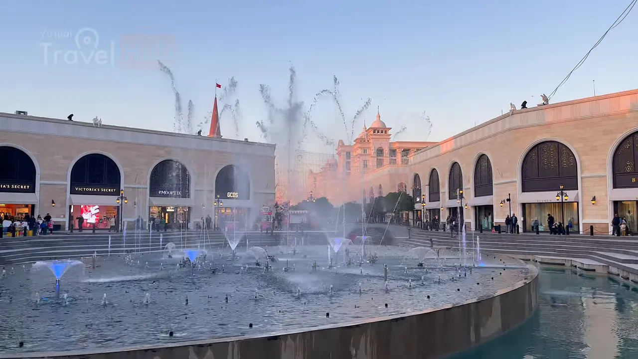 Dusk scene of a circular fountain performing illuminated jets with a warmly lit hotel building behind