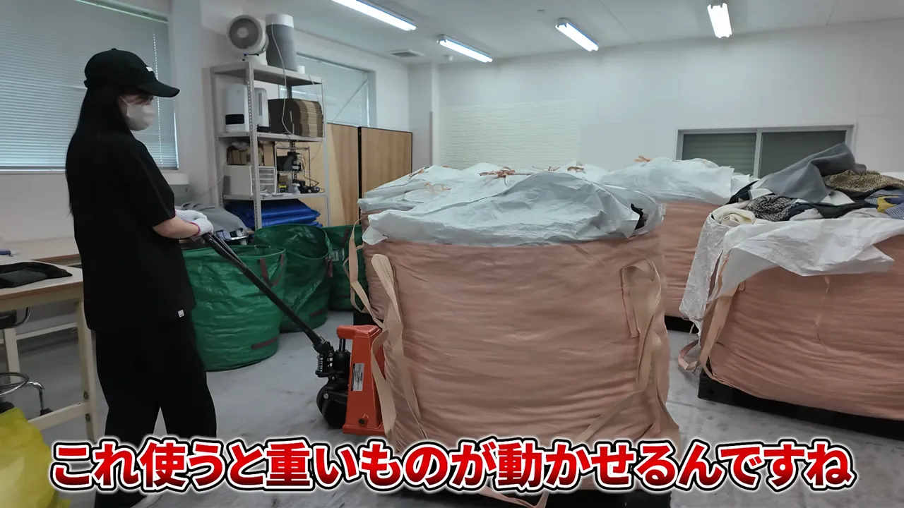 スタッフがオレンジ色のハンドリフトで大きな袋を載せたパレットを動かし、200キロくらいと表示されている様子