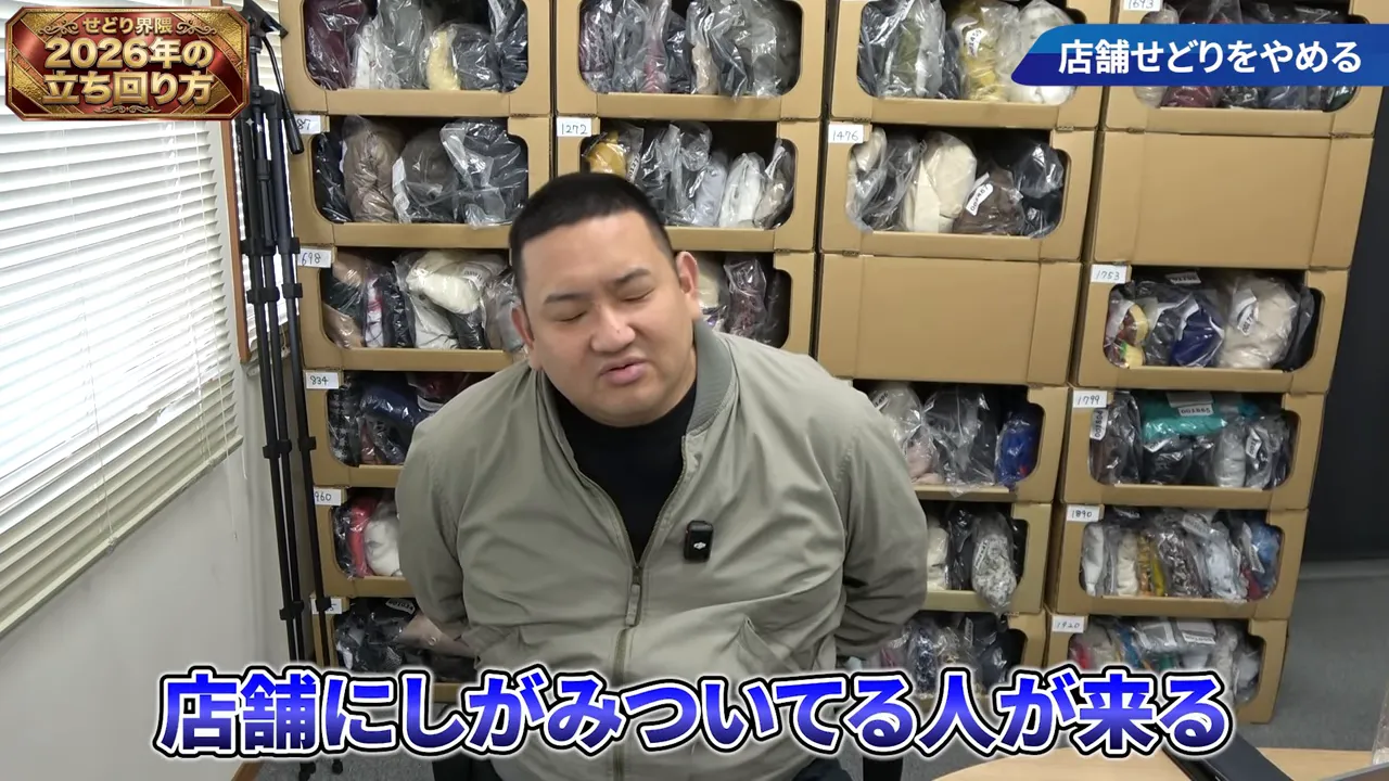 段ボール棚に詰められた包装済み中古商品を背景に座る話者、画面右上に小さな見出し