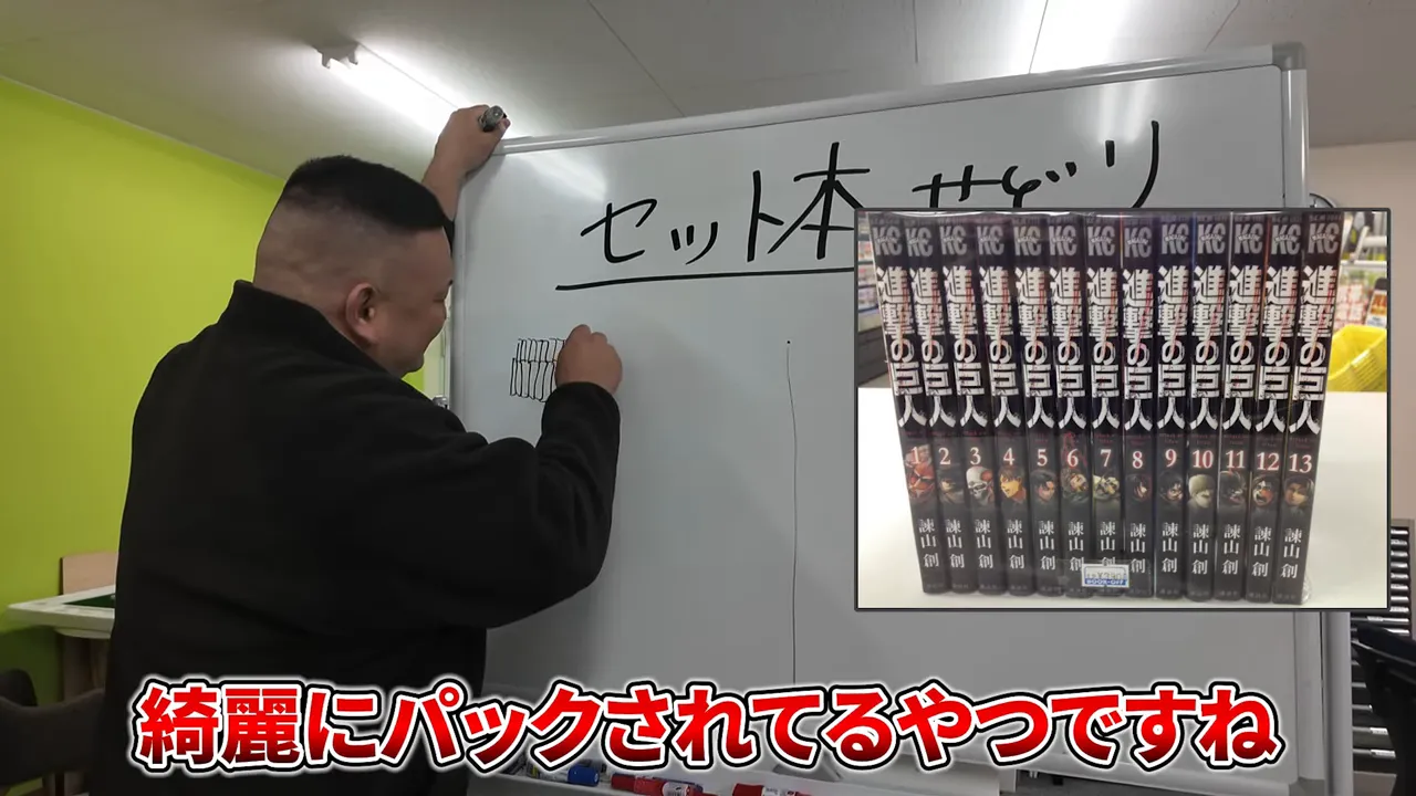 ホワイトボードに説明を書きながら、右上にパックされたマンガ全巻セットの挿し絵が見えるカット