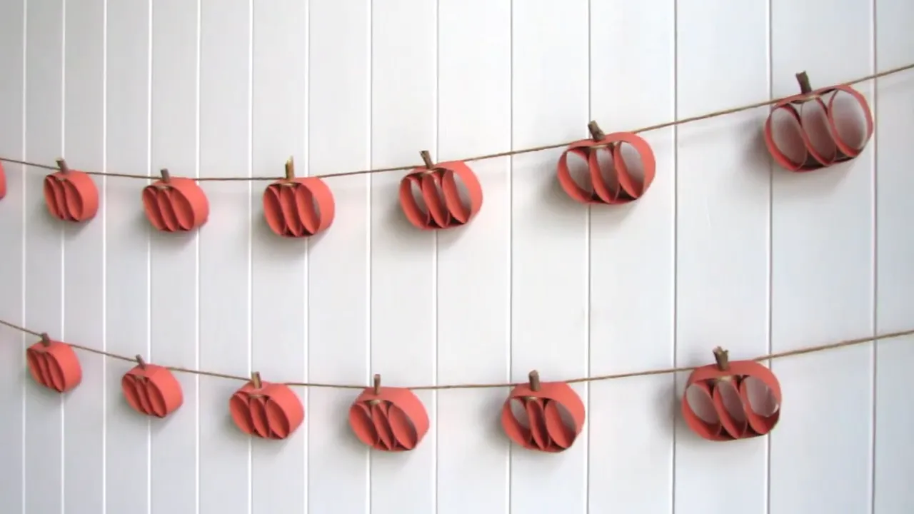 Two rows of red orange paper roll pumpkins on twine against a white wall