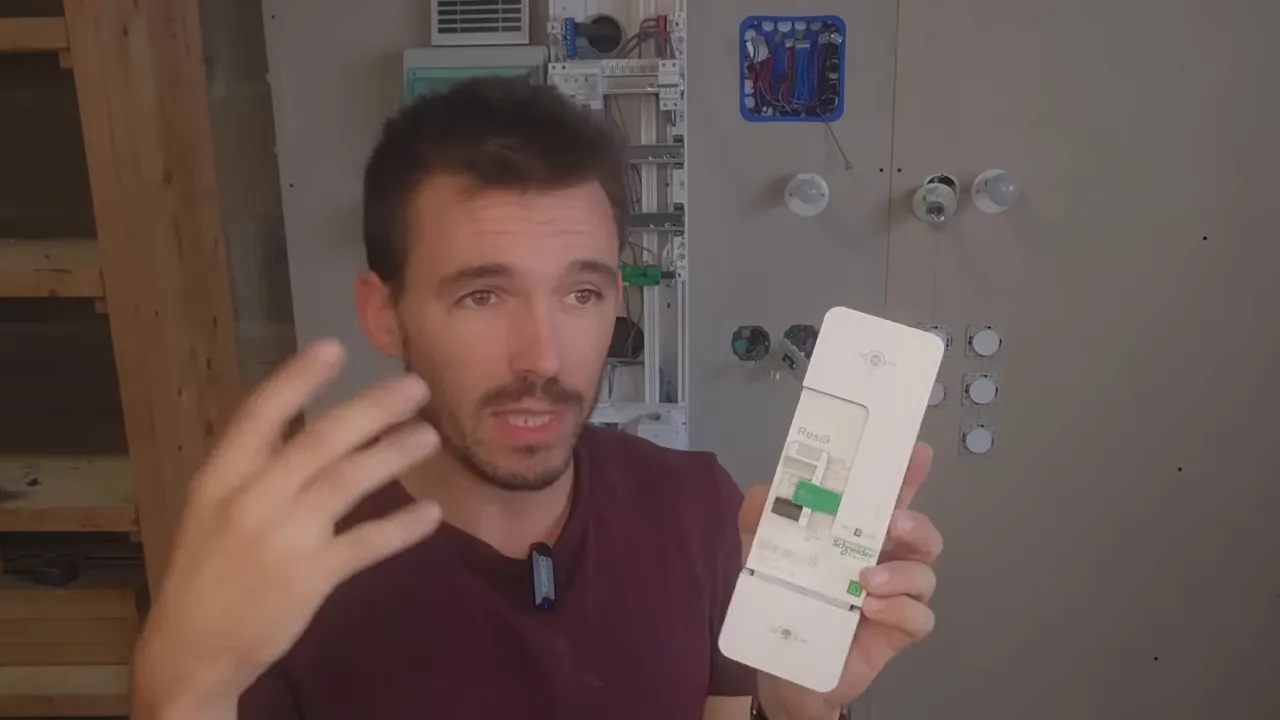 Plan moyen du présentateur expliquant le disjoncteur principal, dispositif blanc bien visible devant un tableau électrique