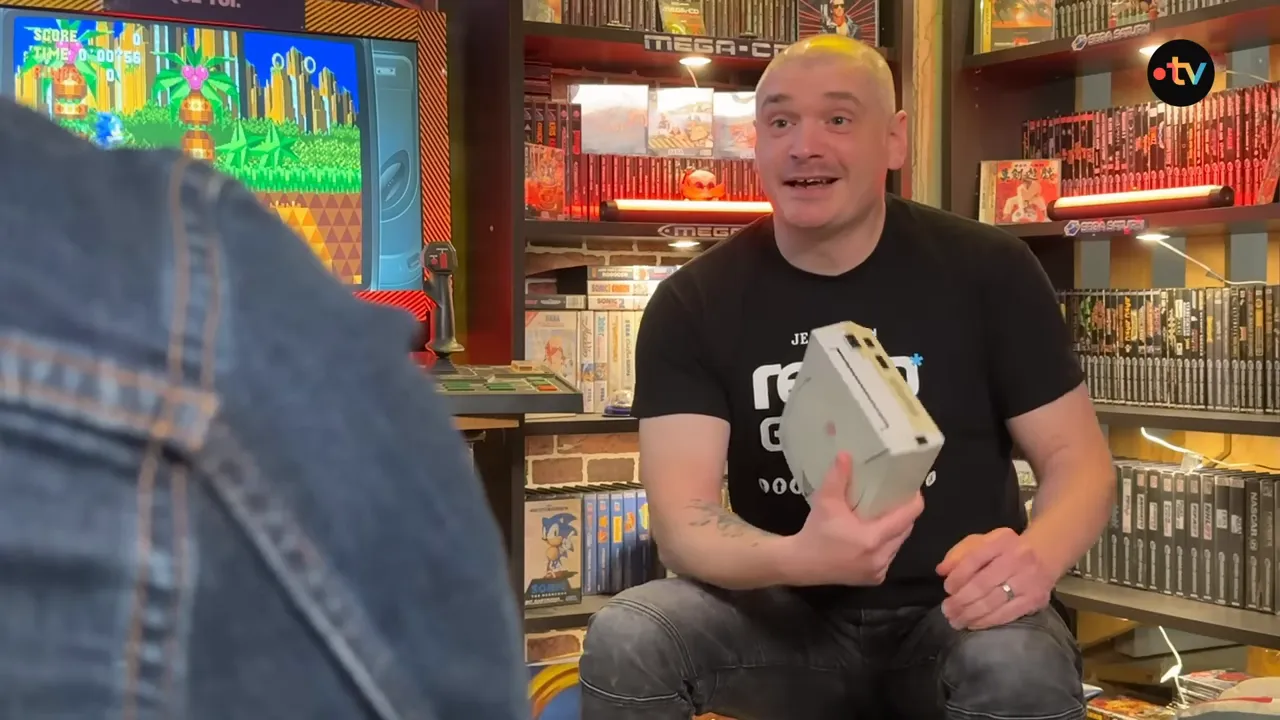 Collectionneur assis tenant une console Dreamcast grise devant des écrans de jeu