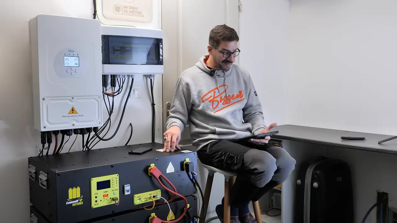 Modules de batteries lithium plug-and-play avec panneaux de contrôle et câbles, à côté d'un onduleur mural, personne assise à droite.