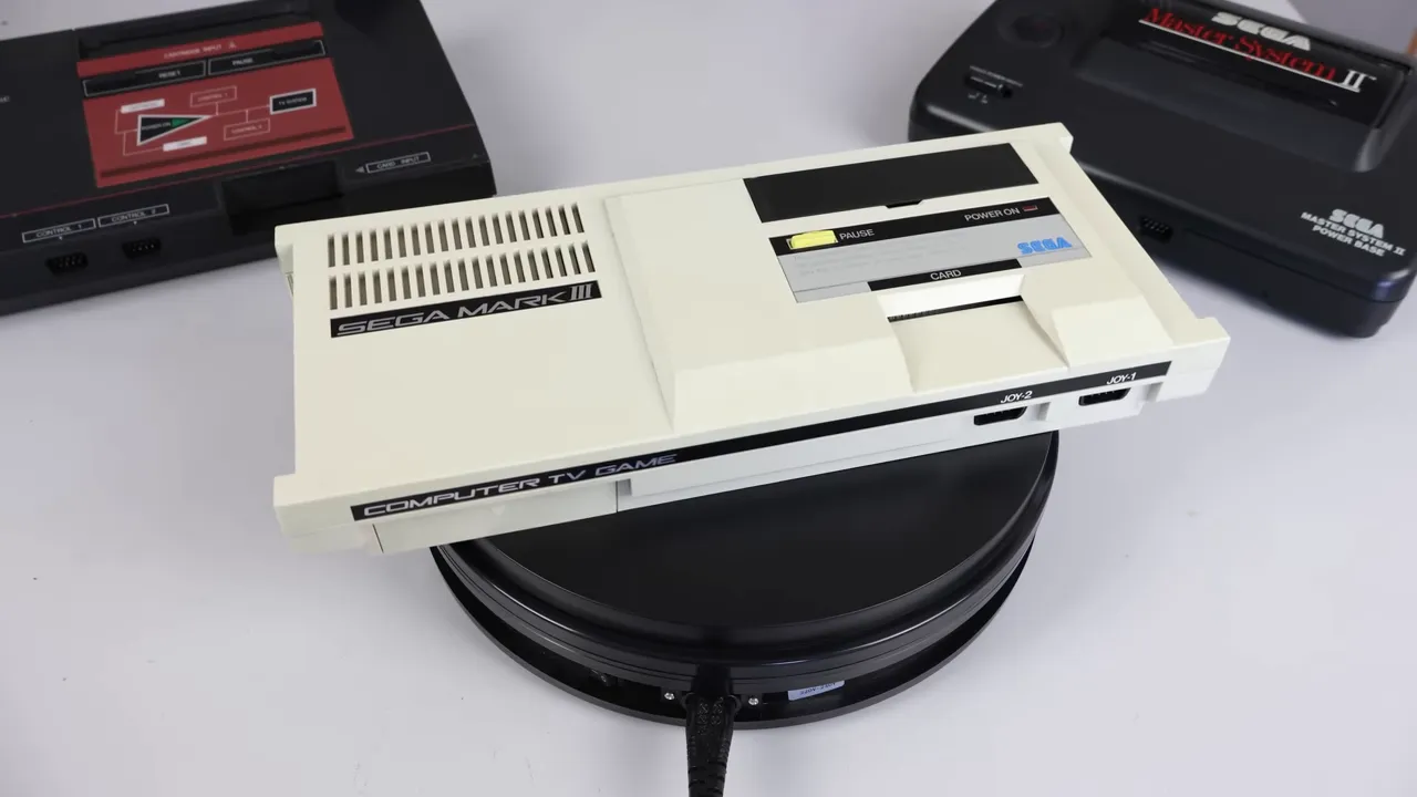 Sega Mark III vue de dessus avec le slot 'CARD' et les prises JOY visibles; Master System et Master System II en arrière-plan pour comparaison