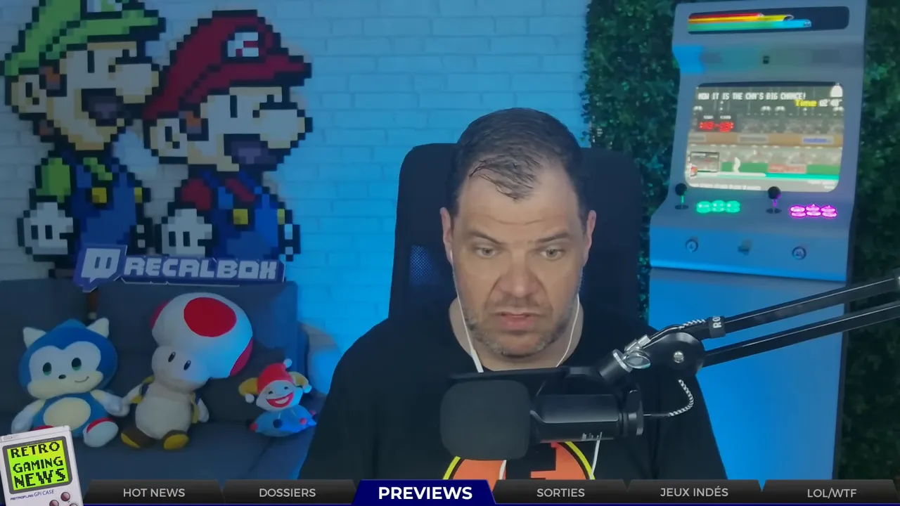 Plan large de l'animateur pendant le segment PREVIEWS, avec une borne d'arcade et des peluches rétro en arrière‑plan ; bandeau PREVIEWS visible en bas.