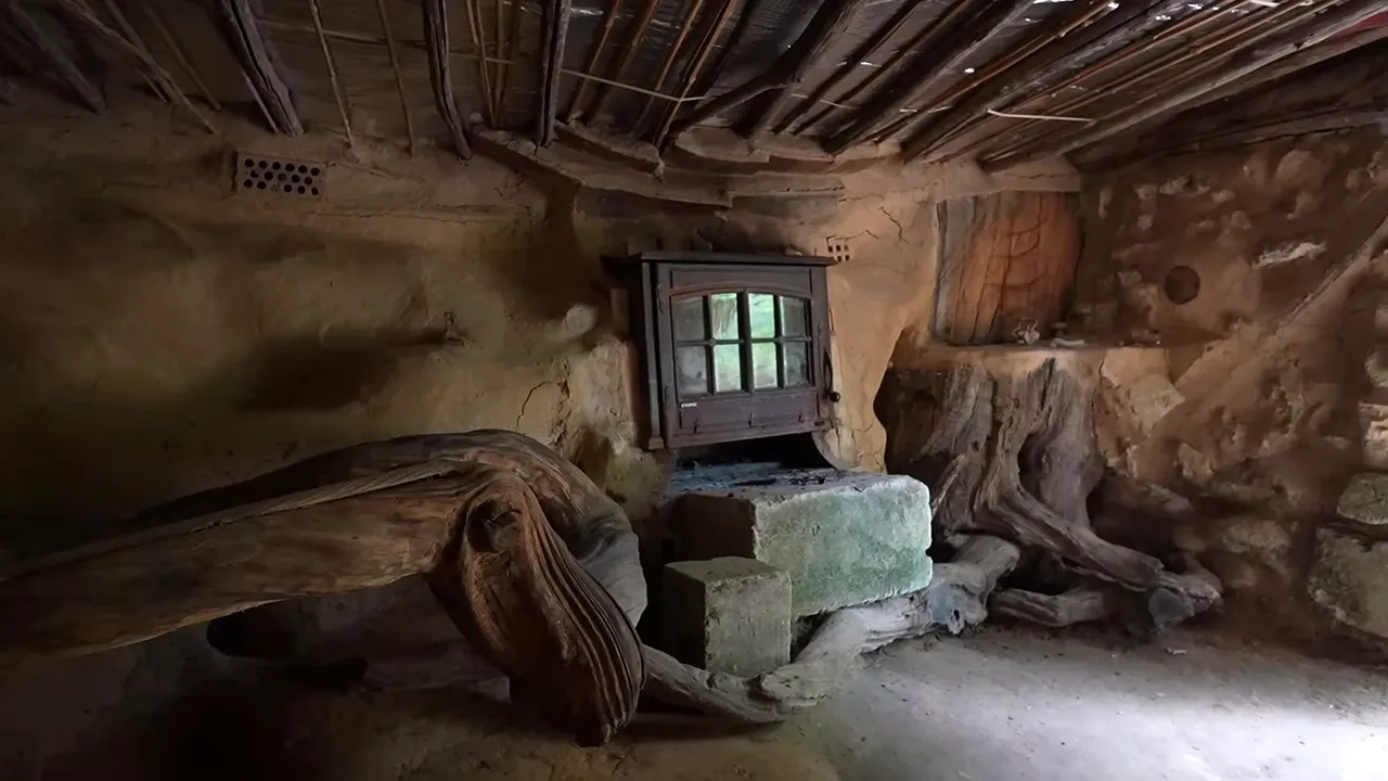 Intérieur de cabane semi-enterrée avec murs en terre, banquettes en bois et poêle