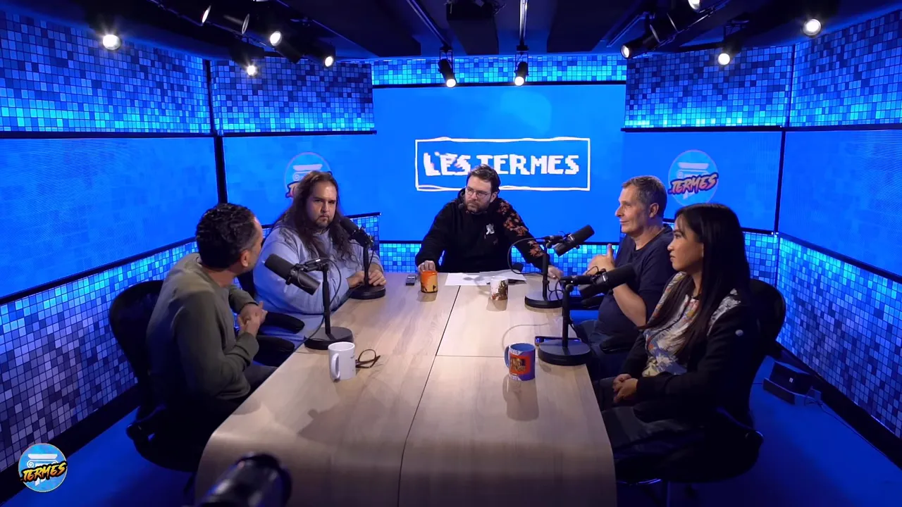 Plan large du studio bleu avec l'équipe assise autour d'une table et le logo de l'émission affiché au centre de l'écran