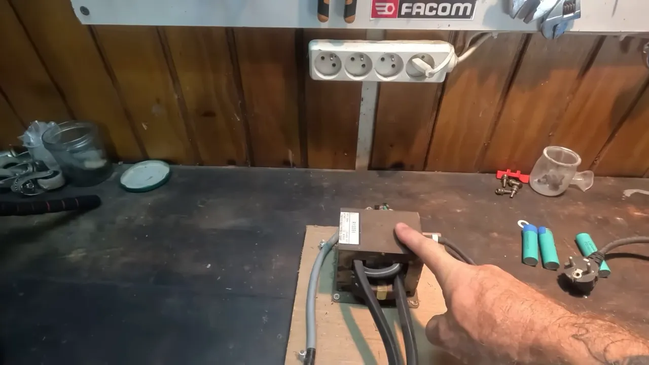 Gros plan d'un transformateur de micro-ondes fixé sur une planche en bois, avec câbles épais et un doigt pointant dessus