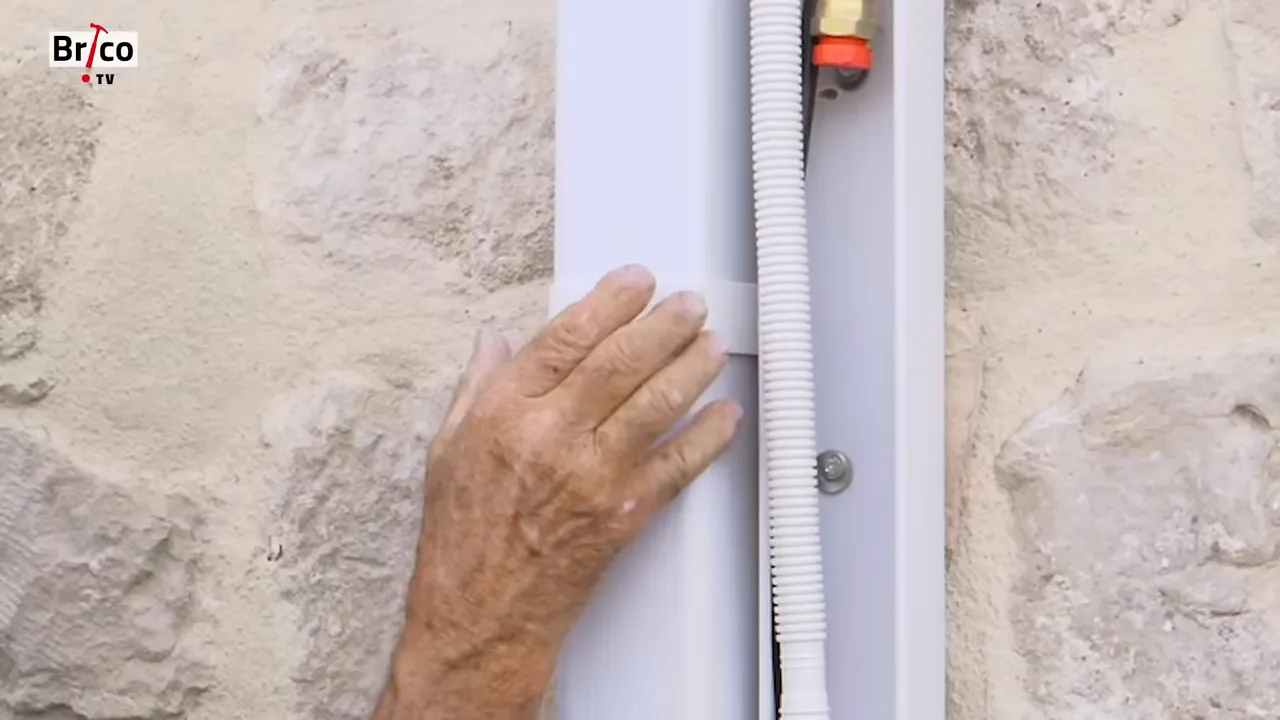 Gros plan sur une goulotte verticale blanche clipsée contre un mur en pierre avec tuyaux et gaine visibles