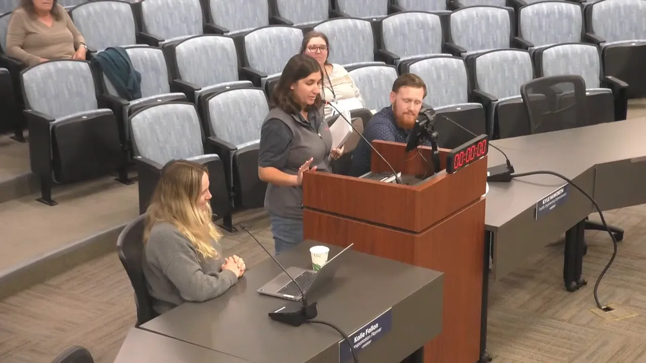 Wider view of a speaker at a podium addressing the Transportation Advisory Board with an attendee at a nearby table and empty audience seats