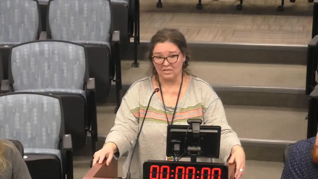 Clear, centered view of a person speaking at a meeting podium with microphone, digital timer, and stepped auditorium seating behind.