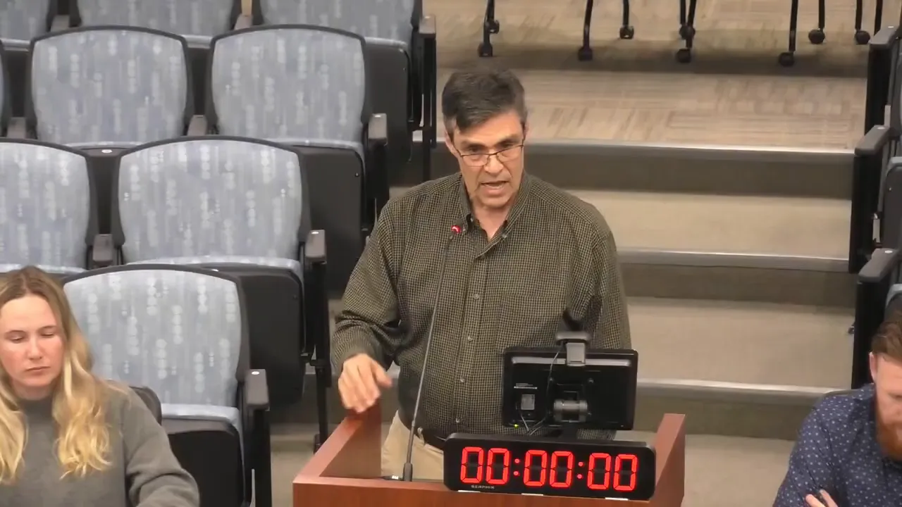 Clear, well-lit view of a person speaking and gesturing at a meeting podium, digital timer visible and auditorium seating behind.