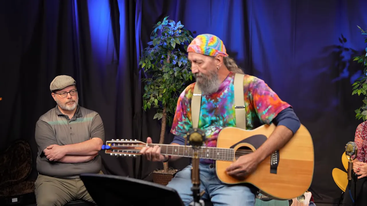 Longmont Song Swap - October 2025 3 Performer strumming an acoustic guitar onstage wearing a colorful bandana, clear and well-lit image.