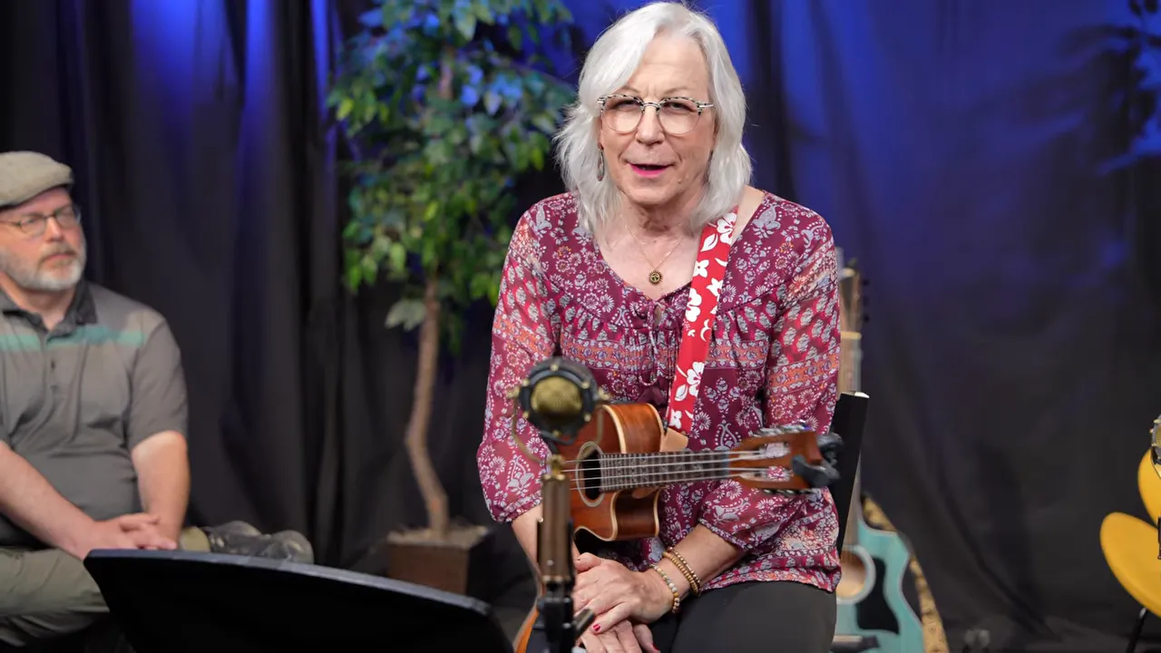 Longmont Song Swap - October 2025 2 seated performer holding a ukulele in front of a microphone on a small stage with plants and black backdrop