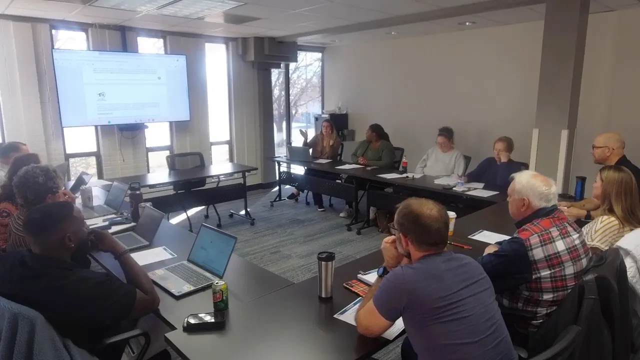Board meeting wide shot with a presenter gesturing and members seated around a table, screen at front
