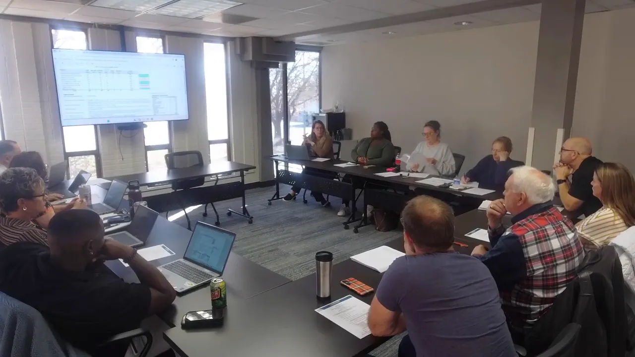 Board members listening to staff presentation in a meeting room with a large screen visible