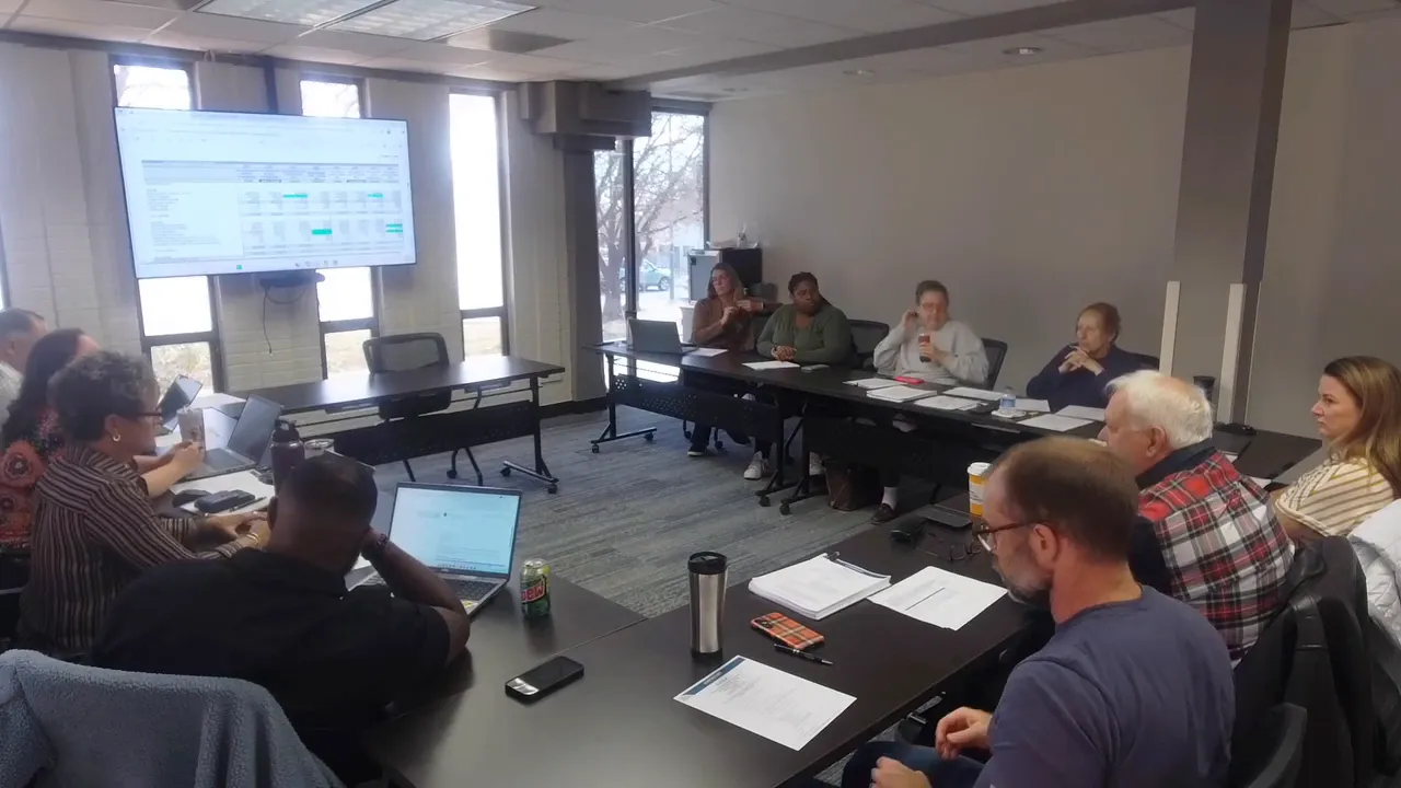 Conference-room shot of the Longmont Housing Authority meeting with a wall-mounted TV displaying a spreadsheet or financial slide and commissioners at a table.